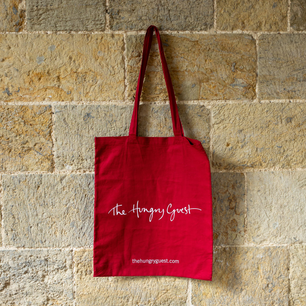 Hungry Guest canvas tote bag in red hanging on a brick wall